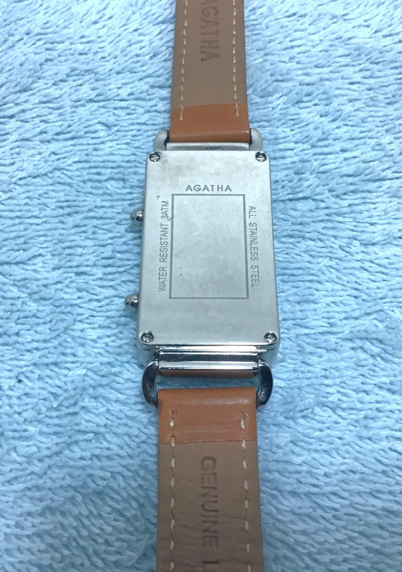 May include: A silver-toned watch with a brown leather band. The watch face has the brand name "AGATHA" and the text "WATER RESISTANT 3ATM" and "ALL STAINLESS STEEL". The band is marked "GENUINE LEATHER".