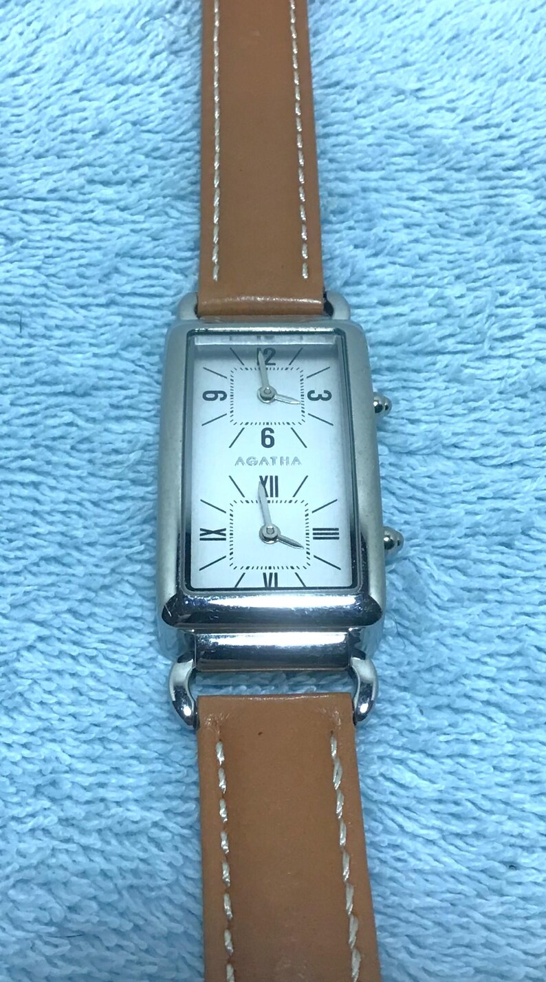 May include: A rectangular silver watch with a white face and brown leather band. The watch face displays the numbers 3, 6, 9, and 12, with the brand name "AGATHA" below the 6. The band is stitched with white thread.