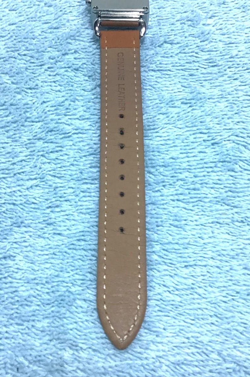 May include: Close-up of a watch with a light brown leather strap. The strap is stitched with white thread and has several holes. The words "GENUINE LEATHER" are printed on the strap.