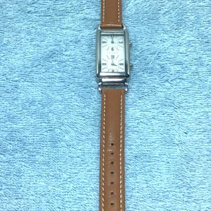 May include: A rectangular wristwatch with a silver-colored face and brown leather band. The watch face displays the numbers 3, 6, 9, and XII. The watch band is stitched with white thread.