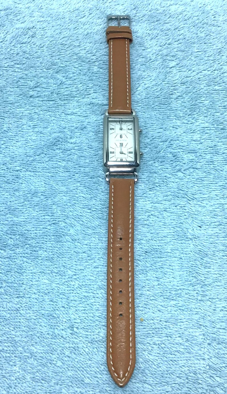 May include: A rectangular wristwatch with a silver-colored face and brown leather band. The watch face displays the numbers 3, 6, 9, and XII. The watch band is stitched with white thread.