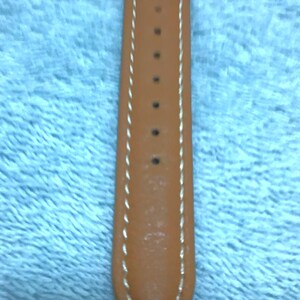 May include: Close-up of a tan leather watch strap with white stitching along the edges. The strap has several holes for adjustment and is tapered at the end. The background is a light blue fabric.