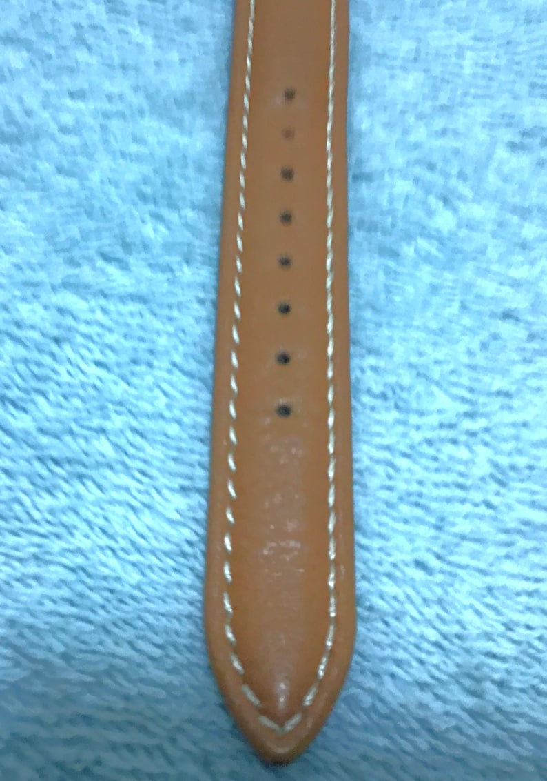 May include: Close-up of a tan leather watch strap with white stitching along the edges. The strap has several holes for adjustment and is tapered at the end. The background is a light blue fabric.