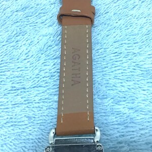 May include: Close-up of an Agatha watch with a brown leather strap. The watch face is rectangular with the brand name Agatha printed on it. The strap is brown with the brand name Agatha printed on it.