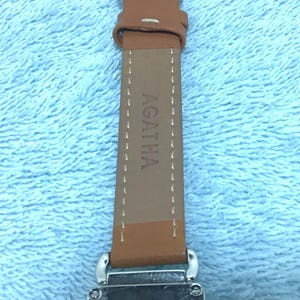 May include: Close-up of an Agatha watch with a brown leather strap. The watch face is rectangular with the brand name Agatha printed on it. The strap is brown with the brand name Agatha printed on it.