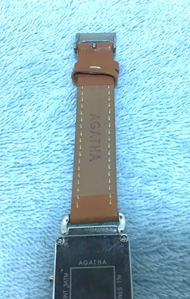 May include: Close-up of an Agatha watch with a brown leather strap. The watch face is rectangular with the brand name Agatha printed on it. The strap is brown with the brand name Agatha printed on it.