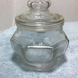 May include: A clear glass jar with a lid. The jar has a hexagonal shape with a rectangular indentation on the front. The lid has a decorative knob. The jar is sitting on a blue surface.