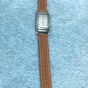 May include: A rectangular silver wristwatch with a brown leather band. The watch face has a white background with black numerals and hands. The band is approximately 0.75 inches wide.