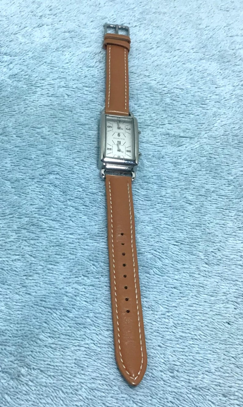 May include: A rectangular silver wristwatch with a brown leather band. The watch face has a white background with black numerals and hands. The band is approximately 0.75 inches wide.
