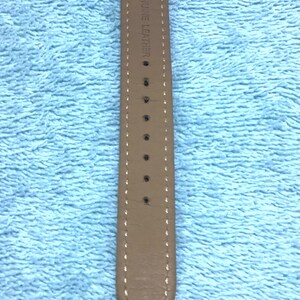 May include: A close-up of a watch with a light brown leather strap. The strap is stitched with white thread and has several holes. The words "GENUINE LEATHER" are printed on the strap.