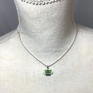 May include: A silver-toned necklace with a small, green, leaf-shaped pendant. The pendant has a dark green outline and a lighter green interior. The word "BUDS" is written in black letters at the bottom of the pendant. The necklace is displayed on a white mannequin.