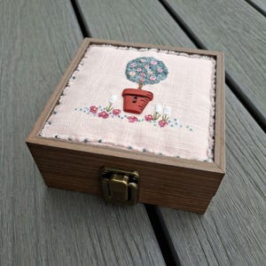May include: A square wooden box with a metal latch, featuring a light pink fabric top embroidered with a potted tree design. The tree has a round top with small pink flowers, a brown button trunk, and a red button pot, surrounded by small floral accents.