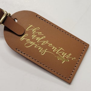 May include: Brown leather luggage tag with a gold strap and the text "The adventure begins" in gold script with a small airplane icon.