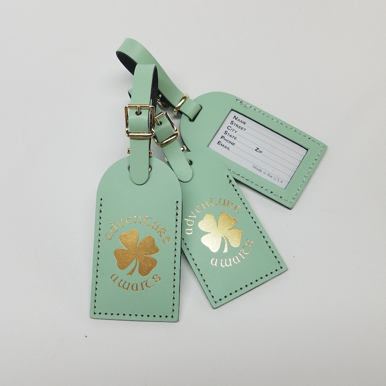 May include: Two green leather luggage tags with gold cloverleaf designs and the text "adventure awaits". One tag has a blank space for writing personal information, including name, street, city, state, zip, phone, and email.