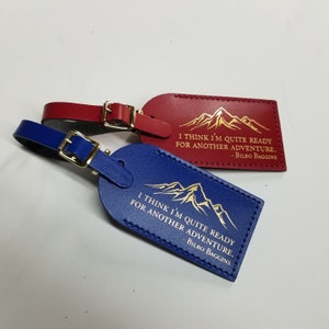 Op de afbeelding: Twee leren bagagelabeltjes met gouden letters. Het rode labeltje zegt "Ik denk dat ik klaar ben voor een nieuw avontuur. - Bilbo Balings". Het blauwe labeltje zegt "Ik denk dat ik klaar ben voor een nieuw avontuur. - Bilbo Balings".