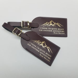 Op de afbeelding: Twee bruine leren bagagelabeltjes met gouden letters. De labels hebben een bergketen afbeelding en de quote "Ik denk dat ik klaar ben voor een nieuw avontuur. - Bilbo Baggins".