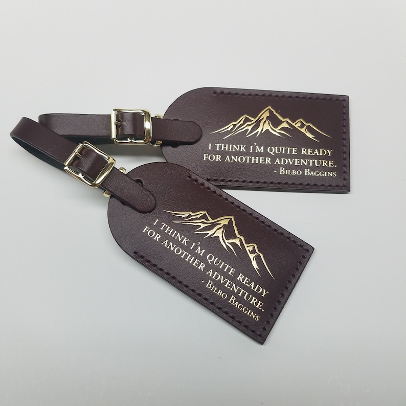 Op de afbeelding: Twee bruine leren bagagelabeltjes met gouden letters. De labels hebben een bergketen afbeelding en de quote "Ik denk dat ik klaar ben voor een nieuw avontuur. - Bilbo Baggins".