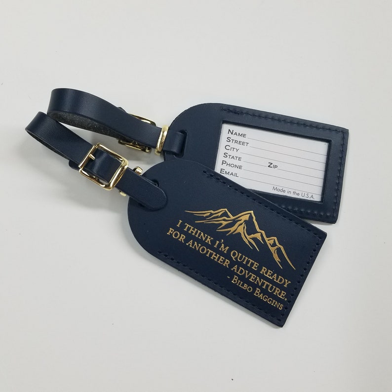 Op de afbeelding: Twee donkerblauwe leren bagagelabeltjes met gouden accenten. E&eacute;n label heeft een bergketenontwerp en de quote "Ik denk dat ik klaar ben voor een nieuw avontuur. - Bilbo Balings". Het andere label heeft een lege ruimte om contactgegevens op te schrijven.