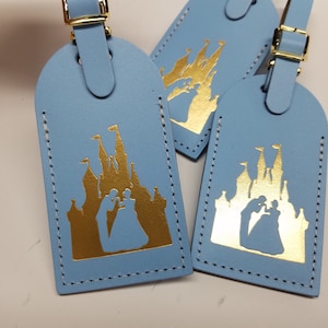May include: Three light blue leather luggage tags with gold stitching. Each tag features a gold silhouette of Cinderella's castle with a couple inside.