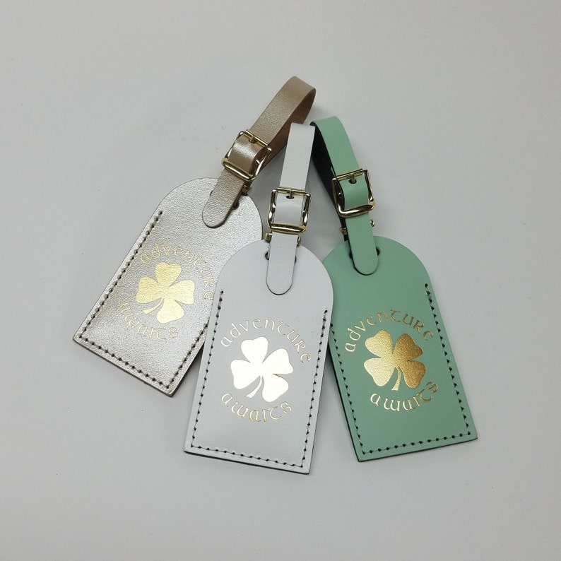 May include: Three leather luggage tags with gold clover designs and the text "Adventure awaits". The tags are in white, light green, and beige.