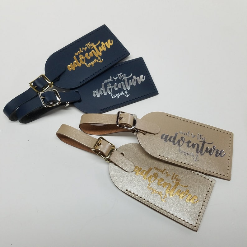 May include: Four leather luggage tags with a gold and silver inscription that reads "and so the adventure begins". Two tags are navy blue and two are beige.