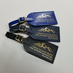 Op de afbeelding: Drie leren bagagelabeltjes met een bergontwerp en de quote "Ik denk dat ik klaar ben voor een nieuw avontuur." van Bilbo Balings. De labeltjes zijn in blauw, donkerblauw en zwart.