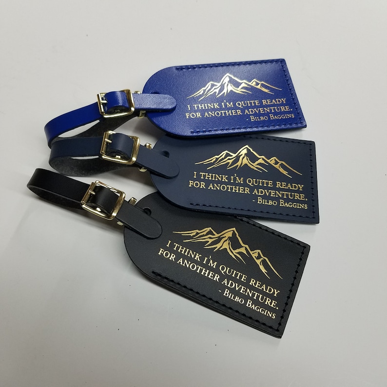 Op de afbeelding: Drie leren bagagelabeltjes met een bergontwerp en de quote "Ik denk dat ik klaar ben voor een nieuw avontuur." van Bilbo Balings. De labeltjes zijn in blauw, donkerblauw en zwart.