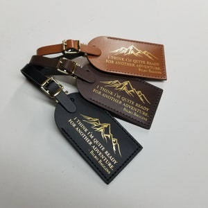 Op de afbeelding: Drie leren bagagelabeltjes met een bergketenontwerp en de quote "Ik denk dat ik helemaal klaar ben voor een nieuw avontuur." van Bilbo Balings. De labeltjes zijn bruin, donkerbruin en zwart.
