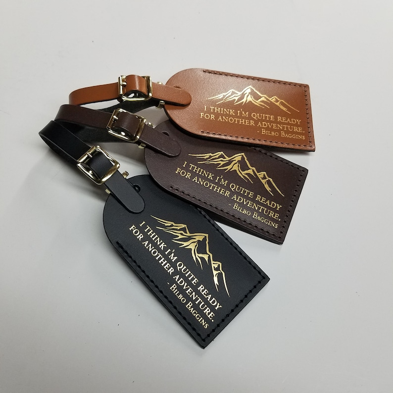 Op de afbeelding: Drie leren bagagelabeltjes met een bergketenontwerp en de quote "Ik denk dat ik helemaal klaar ben voor een nieuw avontuur." van Bilbo Balings. De labeltjes zijn bruin, donkerbruin en zwart.