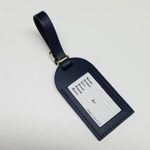 Op de afbeelding: Een donkerblauwe leren bagagelabel met een transparant plastic venster om persoonlijke informatie op te schrijven. Het label heeft gestikte randen en staat onderaan "Made in the U.S.A.".