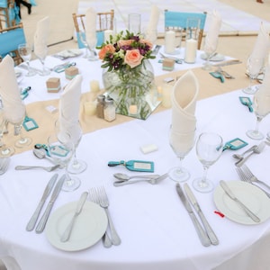 May include: A white tablecloth with a beige runner covers a round table set for a wedding reception. The table is decorated with a centerpiece of pink and peach flowers in a clear glass vase. White napkins are folded and placed on each plate, and there are clear glass wine glasses and silverware on the table.