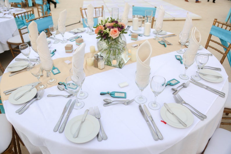 May include: A white tablecloth with a beige runner covers a round table set for a wedding reception. The table is decorated with a centerpiece of pink and peach flowers in a clear glass vase. White napkins are folded and placed on each plate, and there are clear glass wine glasses and silverware on the table.