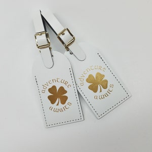 May include: Two white leather luggage tags with gold stitching and a gold four-leaf clover design. The text "adventure awaits" is printed on each tag.