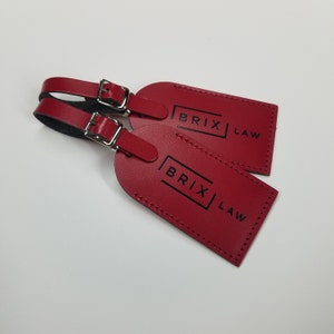 May include: Two rectangular red luggage tags with rounded ends, featuring a silver metal buckle strap and the black text BRIX LAW printed on the front. The tags have visible stitching along the edges and a smooth, consistent red finish.