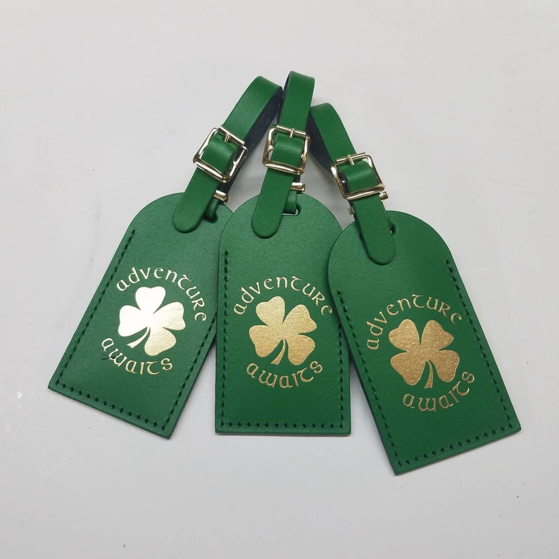 May include: Three green leather luggage tags with gold lettering that reads "adventure awaits" and a gold four-leaf clover design.