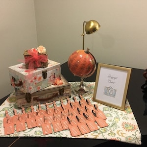 May include: A table setting for a travel-themed party with a globe, luggage-shaped boxes, and a sign that says "Passport to Fun". There are also small cards with pink and white accents.