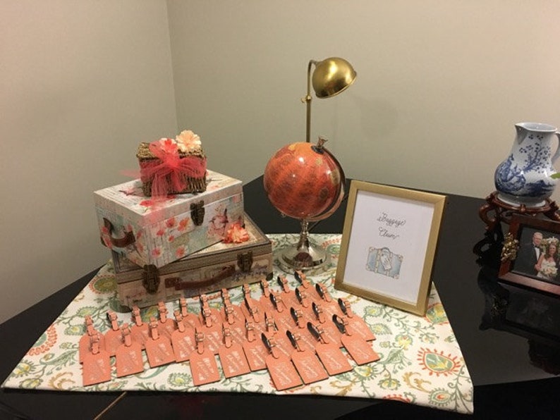 May include: A table setting for a travel-themed party with a globe, luggage-shaped boxes, and a sign that says "Passport to Fun". There are also small cards with pink and white accents.