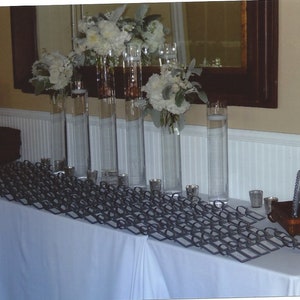 May include: A white tablecloth with a black metal frame holding place cards. There are several tall glass vases filled with white flowers and greenery. A small wooden box with a silver frame is on the table.
