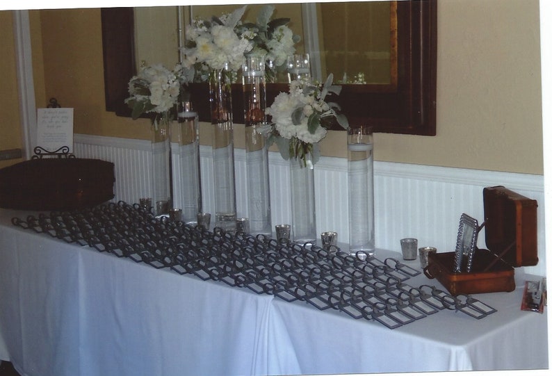 May include: A white tablecloth with a black metal frame holding place cards. There are several tall glass vases filled with white flowers and greenery. A small wooden box with a silver frame is on the table.