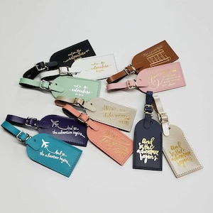 May include: A collection of 12 leather luggage tags with gold lettering. The tags feature various travel-themed quotes, such as "And so the adventure begins" and "Just the beginning".