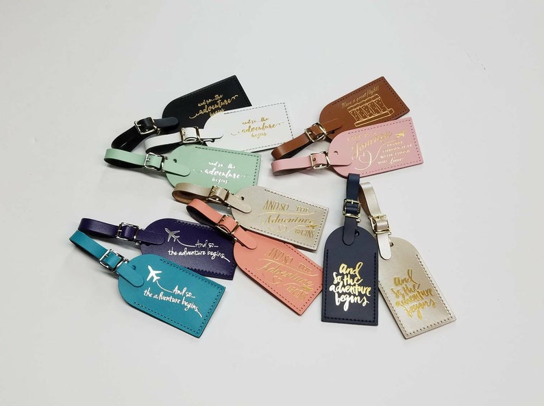 May include: A collection of 12 leather luggage tags with gold lettering. The tags feature various travel-themed quotes, such as "And so the adventure begins" and "Just the beginning".