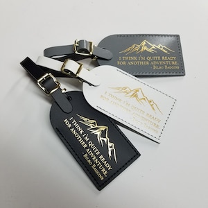 Op de afbeelding: Drie leren bagagelabeltjes met een bergontwerp en de quote "Ik denk dat ik klaar ben voor een nieuw avontuur. - Bilbo Balings" in gouden letters. De labeltjes zijn zwart, wit en grijs.