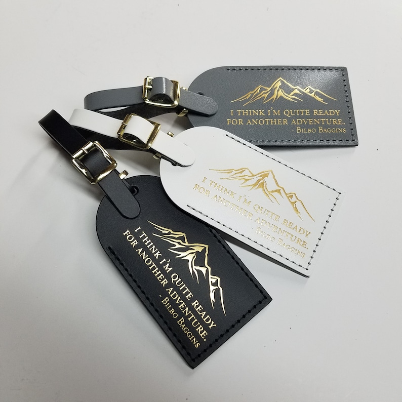 Op de afbeelding: Drie leren bagagelabeltjes met een bergontwerp en de quote "Ik denk dat ik klaar ben voor een nieuw avontuur. - Bilbo Balings" in gouden letters. De labeltjes zijn zwart, wit en grijs.