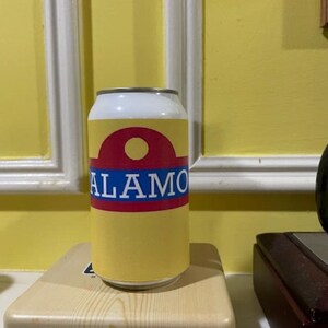 May include: A yellow and white aluminum can with the word "ALAMO" in blue letters. The can has a red and white design at the top and is sitting on a light-colored box.
