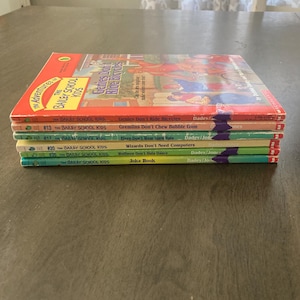 May include: A stack of children's books from "The Bailey School Kids" series. The books have colorful covers with titles like "Genies Don't Ride Bicycles" and "Gremlins Don't Chew Bubble Gum". The books are stacked on a wooden table.