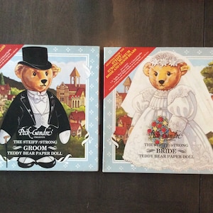 May include: Two vintage Steiff/Strong Teddy Bear paper doll boxes. One features a groom bear in a tuxedo and top hat, the other a bride bear in a wedding dress and veil. Both boxes have a light blue border and text.
