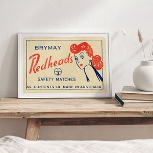 May include: A vintage framed print of a matchbox label for Brymay Redheads Safety Matches. The label features a cartoon illustration of a woman with red hair looking over her shoulder. The text on the label reads "Brymay Redheads Safety Matches Av. Contents 50 Made in Australia."