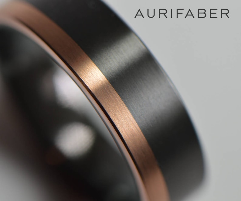 May include: A black and rose gold wedding band with a brushed finish. The band has a thin rose gold inlay around the edge. The band is made by Aurifaber.