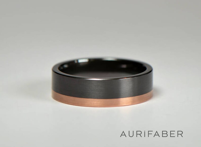 May include: A black and rose gold wedding band with a brushed finish. The band has a thin rose gold stripe around the edge.