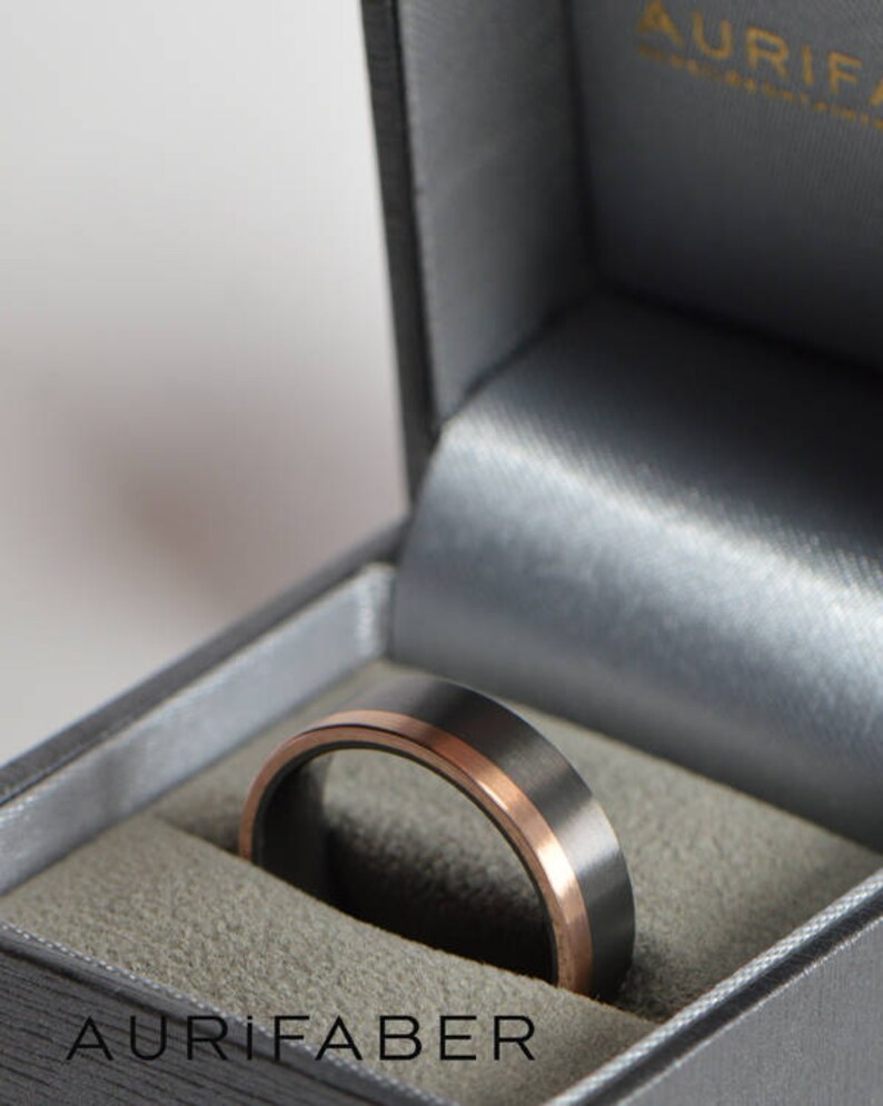 May include: A black and rose gold wedding band in a gray velvet ring box. The box is labeled "Aurifaber".